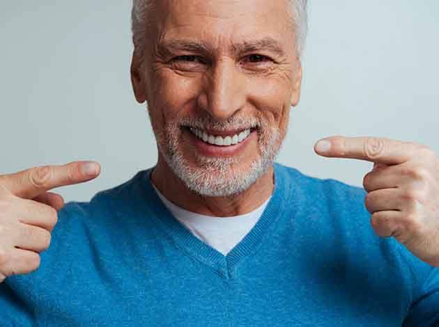 Man in blue sweater with white beard pointing to his smile with his pointer fingers